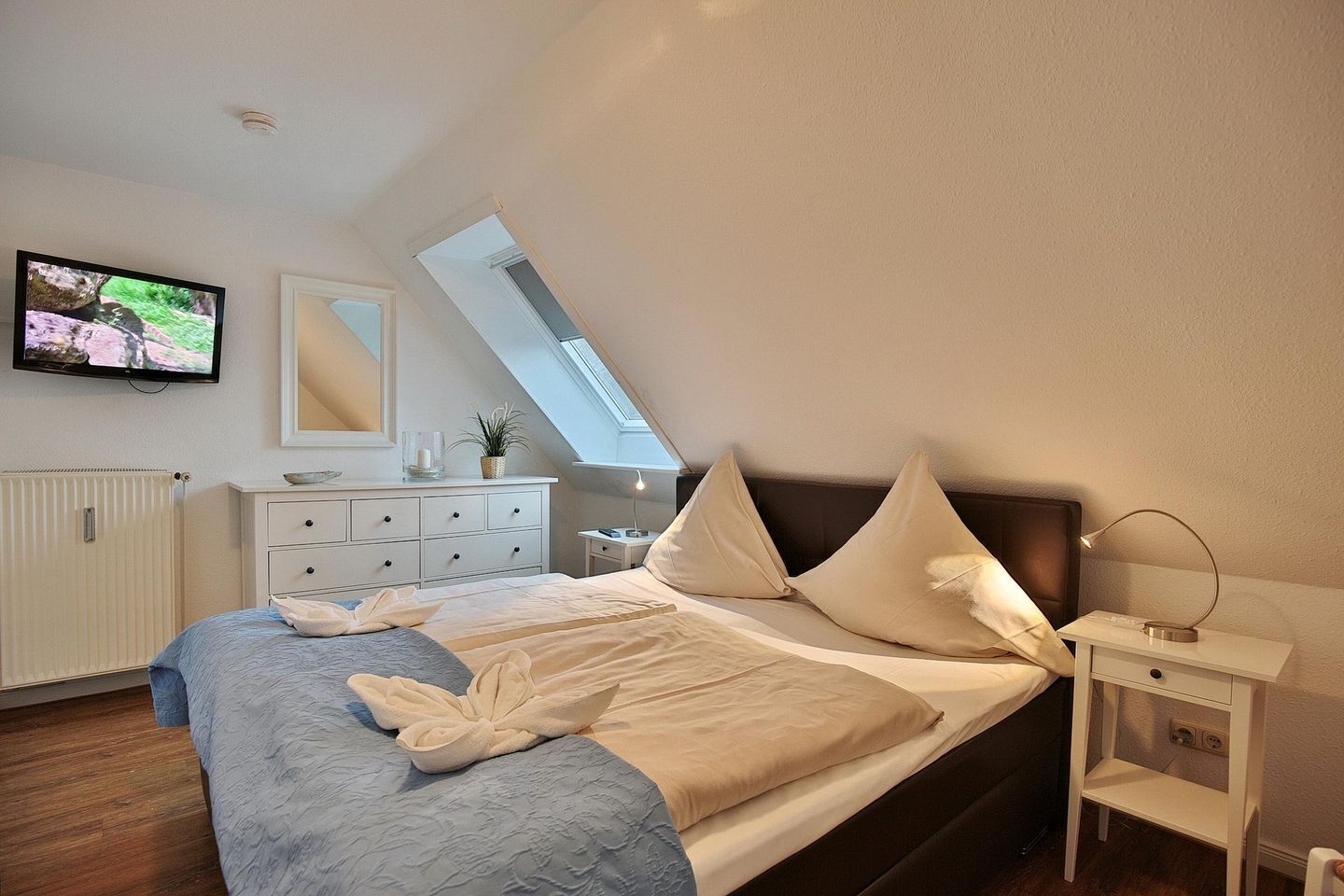 A bedroom with a bed, a bedside table, a lamp, a TV, a dresser, and a skylight.