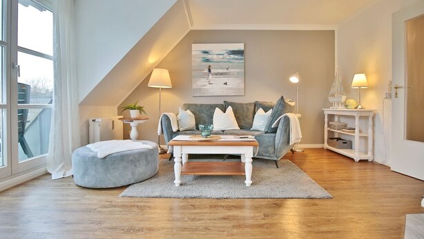A cozy living room with a grey sofa, a white coffee table, and a grey pouf. There is a painting on the wall, a lamp on a side table, and a window with curtains. - Am Elchgrund Apartment 12 - Im Brook 36-38 (Timmendorfer Strand)