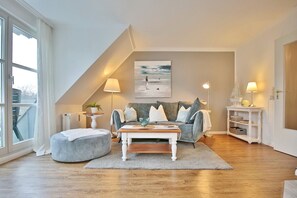 Un salon chaleureux avec un canapé gris, une table basse blanche et un pouf gris. Un tableau est accroché au mur, une lampe se trouve sur une table d’appoint, et une fenêtre est ornée de rideaux.