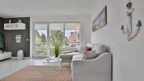 Een moderne woonkamer met een grijze bank, een ronde salontafel en een groot raam met uitzicht op een gebouw en bomen.