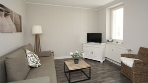 Una sala de estar con un sofá, una mesa de centro, un televisor sobre un mueble, una lámpara y una silla de mimbre.