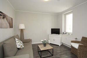 Een woonkamer met een bank, een salontafel, een televisie op een kast, een lamp en een rieten stoel.