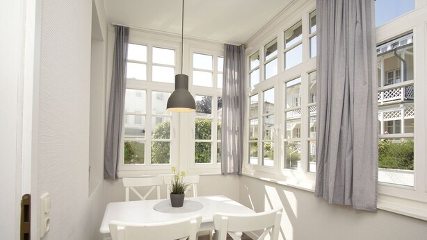 Un comedor con una mesa y sillas blancas, una lámpara colgante y ventanas grandes con cortinas.