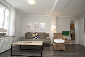 Ein Wohnzimmer mit einer Couch, einem Couchtisch, einem Korbstuhl und einer Lampe. An den Wänden hängen Bilder in Rahmen und links befindet sich eine Heizung.