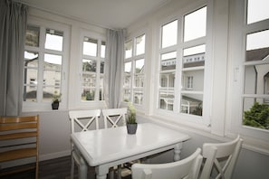 Un comedor con una mesa y sillas blancas, ventanas amplias con cortinas y plantas en macetas sobre los alféizares.
