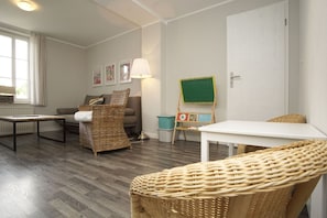 Ein gemütliches Wohnzimmer mit einer Couch, einem Korbsessel, einem kleinen Tisch, einer Lampe und einer grünen Staffelei.