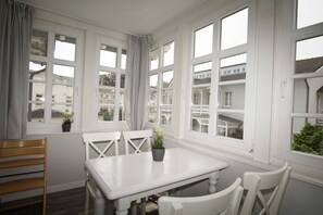 Un coin repas avec une table et des chaises blanches, de grandes fenêtres avec des rideaux et des plantes en pot posées sur les rebords de fenêtre.