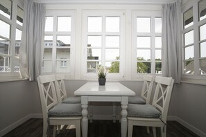 A white table with two chairs in a bay window setting, a potted plant on the table, and checkered curtains on the windows.