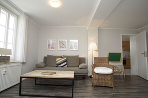 A living room with a sofa, a coffee table, a wicker chair, and a lamp. There are framed pictures on the wall and a radiator on the left. - Vacation apartment in spa style with conservatory - Villa Maria FeWo Marianne 35 - (Göhren)