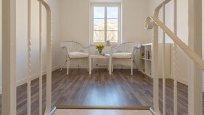 Quarto com chão de madeira, duas cadeiras de vime, uma mesa pequena com um vaso de flores e uma estante de livros.