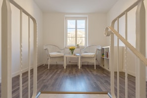 Une pièce avec un plancher en bois, deux fauteuils en osier, une petite table sur laquelle est posé un vase de fleurs et une bibliothèque.