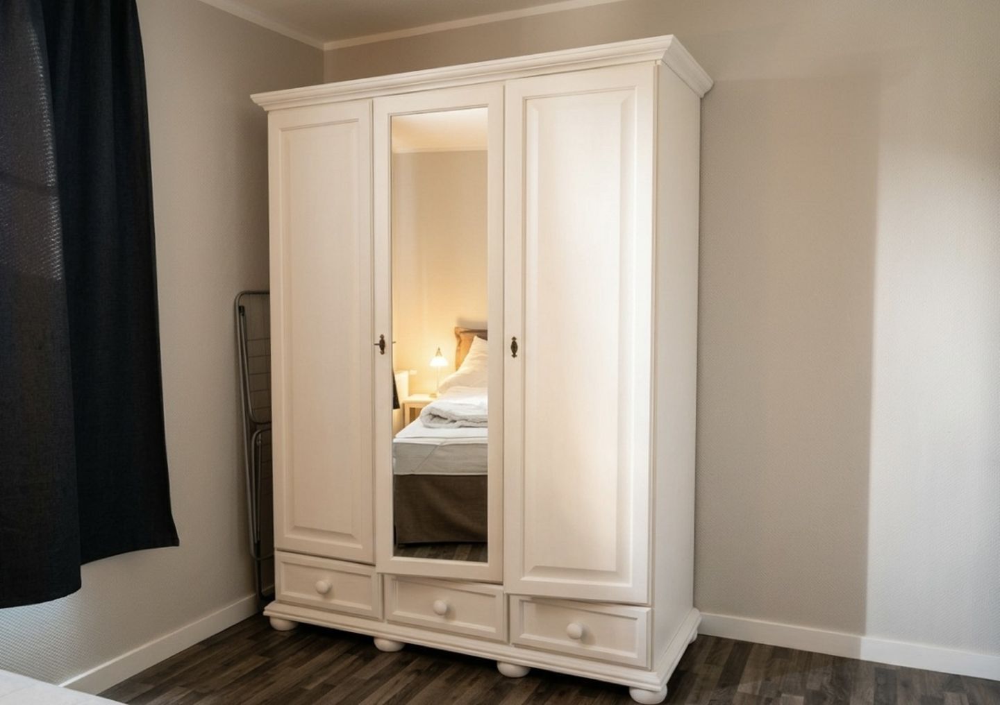 A white wardrobe with a mirror, a bed with a white duvet, and a black curtain.
