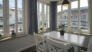 Quarto com uma mesa e cadeiras brancas, grandes janelas com cortinas, luminária pendente e vista para edifícios.