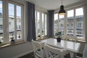A room with a white table and chairs, large windows with curtains, a hanging light fixture, and a view of buildings outside. - Bathroom-style vacation apartment with conservatory - Villa Maria FeWo Maja 36 - Villa (Göhren)