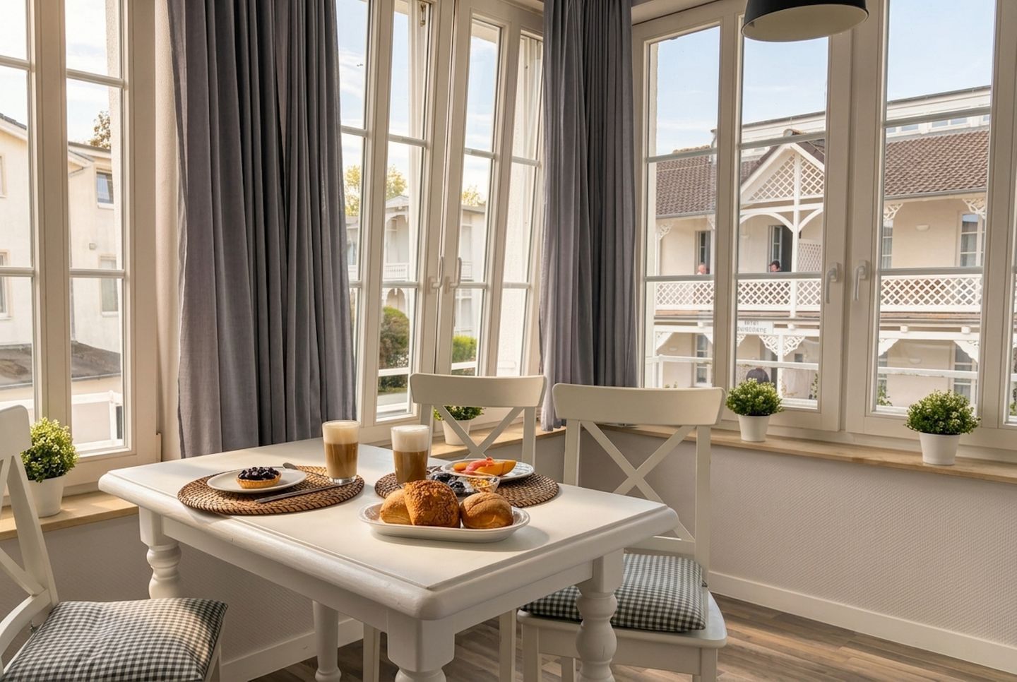 A dining area with a table set for breakfast, including pastries and beverages, with large windows and curtains.