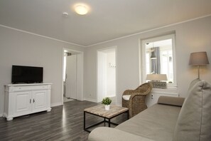 Una sala moderna con un televisor de pantalla plana sobre un mueble blanco, un sofá gris, un sillón de mimbre, una mesita de centro de madera y una lámpara.