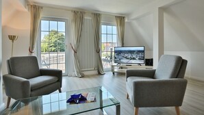 Un salón moderno con dos sillones grises, una mesa de centro de vidrio, un televisor de pantalla plana y vistas a un paisaje urbano a través de amplios ventanales.