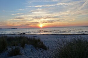 A beach at sunset with the sun setting over the ocean, sand dunes, and sparse vegetation in the foreground. - Vacation apartment in a quiet location and with balcony - Ferienhaus Solamar FeWo OG - (Breege)