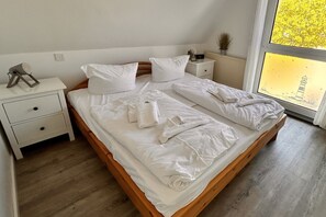 A neatly made bed with white linens and towels, a bedside table with a lamp, and a view of a tree through a window.