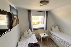 Una habitación pequeña y ordenada con dos camas individuales, una mesita, un televisor montado en la pared y una ventana con cortinas.