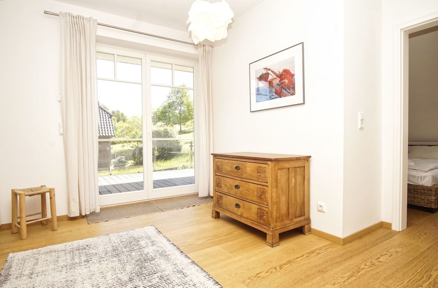 A room with a wooden dresser, a small stool, a rug, a sliding glass door, and a framed picture on the wall.