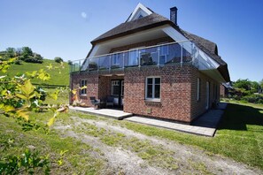 Une maison à deux étages dotée d’un balcon en verre, de murs en briques et d’un toit de chaume.