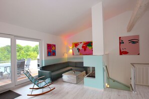 A modern living room with a sectional sofa, a rocking chair, and a fireplace. There are large windows and a view of greenery outside. - Close to the beach, balcony, ocean view, sauna, free adventure pool - Palstek Beach House - (Göhren)