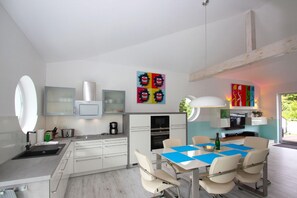 A modern kitchen with white cabinets, a dining table with blue placemats, and a large window. - Close to the beach, balcony, ocean view, sauna, free adventure pool - Palstek Beach House - (Göhren)