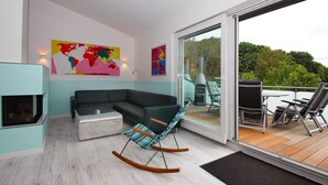 Un salon moderne avec un canapé, un fauteuil à bascule et une cheminée. Il y a une grande fenêtre et une porte qui donne sur une terrasse extérieure équipée de chaises et d’un barbecue.