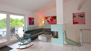 Un salon moderne avec un canapé d’angle, un fauteuil à bascule et une cheminée. De grandes fenêtres offrent une vue sur la verdure environnante.