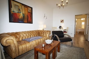 Una sala de estar con un sofá de cuero marrón, una mesa de centro de madera y un comedor con una mesa y sillas blancas.