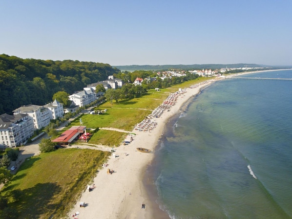 An aerial view of a coastal area with a beach, buildings, and a forested hill.