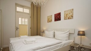 Una cama bien hecha con ropa blanca, una araña y tres cuadros en la pared.