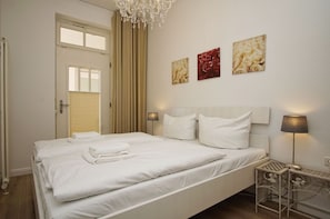 A neatly made bed with white linens, a chandelier, and three framed pictures on the wall.