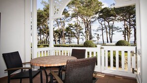 Un patio cubierto con una mesa redonda y sillas de mimbre, con vistas a un área ajardinada con árboles y el mar.