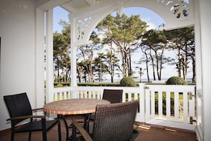 A covered patio with a round table and wicker chairs, overlooking a landscaped area with trees and a view of the sea.