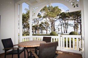 Un patio cubierto con una mesa redonda y sillas de mimbre, con vistas a un área ajardinada con árboles y el mar.