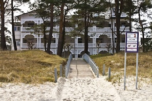 A pathway leading to a white building with balconies, a sign indicating a toilet and textile strand, and a warning about dogs.