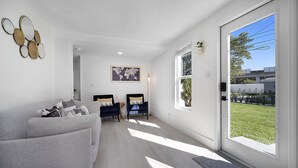Una sala de estar moderna con un sofá gris, dos sillones, un cuadro de un mapa del mundo y un ventanal con vistas a un césped verde y edificios.