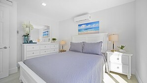 Un dormitorio con cama, cómoda, mesita de noche, espejo, un cuadro de una playa y un equipo de aire acondicionado.