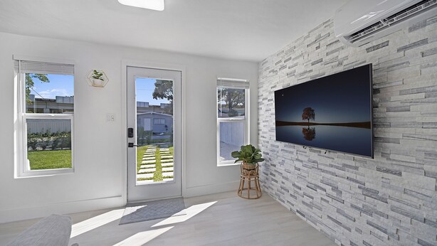Una sala de estar moderna con un televisor grande montado en una pared texturizada, dos ventanas que dan a un jardín y una puerta que da al exterior.