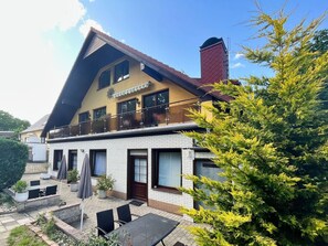 Una casa de dos pisos con un balcón, un patio con mobiliario exterior y un árbol grande al frente.