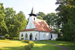 A church with a steeple, arched windows, and a red roof, surrounded by greenery.