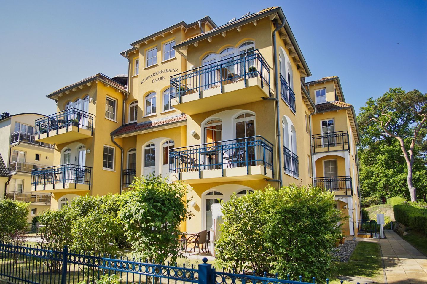 A multi-story yellow building with balconies, a blue fence, and greenery in front.