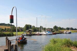 A calm river with boats docked at a pier, surrounded by greenery and a small building.