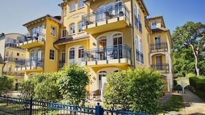 A multi-story yellow building with balconies, a blue fence, and greenery in front. - Kurparkresidenz - close to the beach, free use of the AHOI adventure pool and sauna - (Baabe)