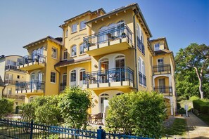 A multi-story yellow building with balconies, a blue fence, and greenery in front.