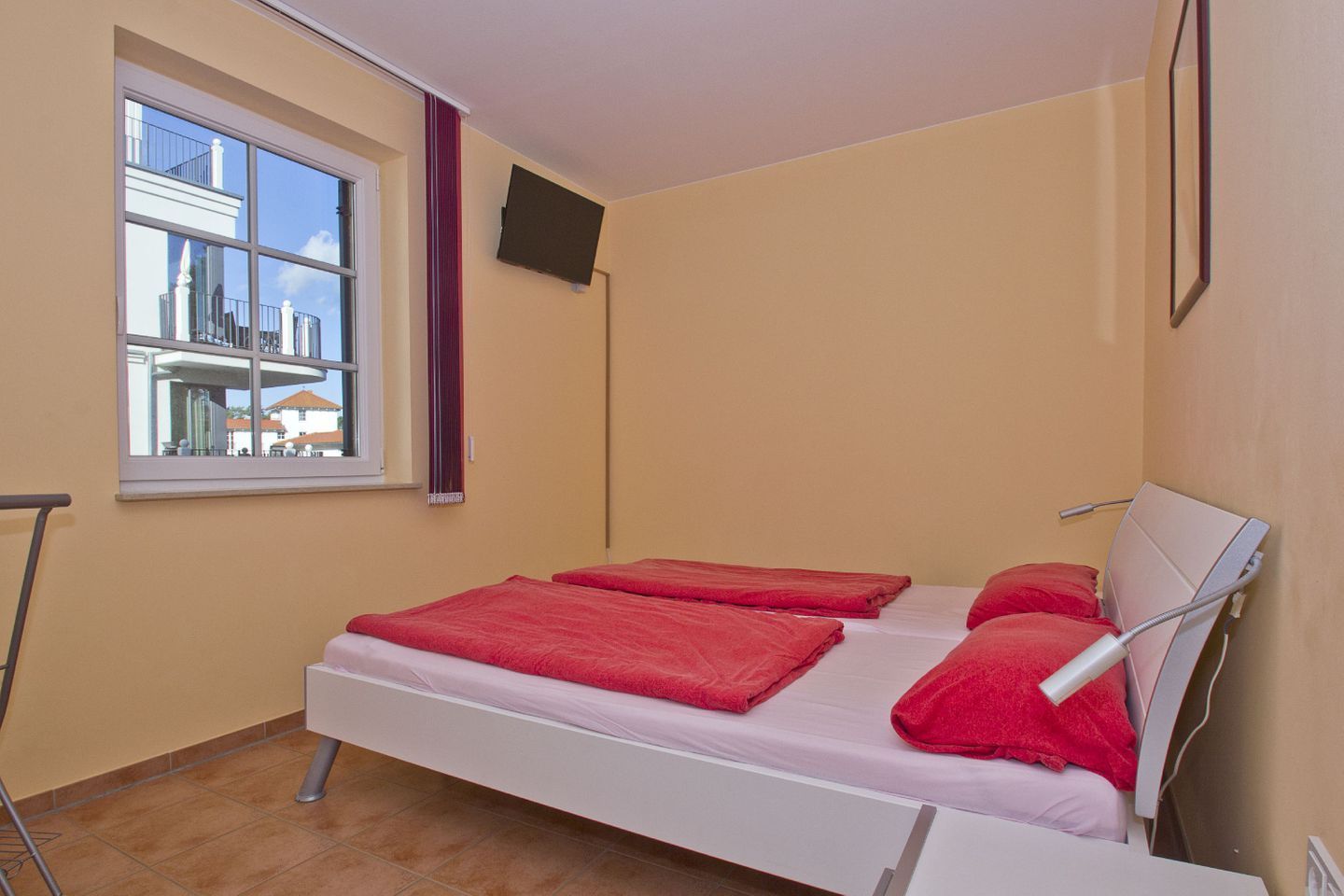 Une petite chambre avec un lit simple, une fenêtre, un téléviseur à écran plat et un lit recouvert de draps rouges.