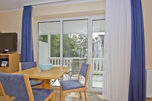 Ein Zimmer mit einem Holztisch, zwei blauen Stühlen, einer blauen Schüssel, einem Fernseher auf einem Standfuß und einer Glastür zu einem Balkon mit weißer Brüstung.