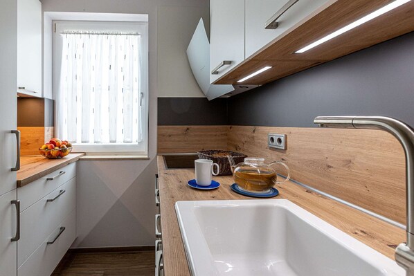 A compact kitchen with a sink, a window with blinds, a bowl of fruit, and a tea setup. - Villa Böck F-1105 - Villa Böck F-1105 An der Düne - Arkonablick View (Glowe)